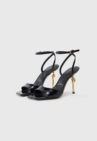 Sandalias de tacón alto de charol negro con punta cuadrada, tiras en el tobillo y acentos de hardware retorcido en tono dorado en el tacón.