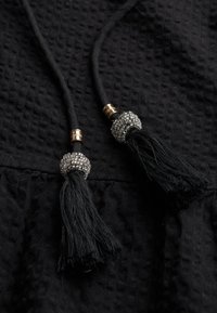 Tela negra con un patrón texturizado que presenta dos borlas negras en los extremos de los cordones, acentuada con herrajes de plata y oro con cuentas.