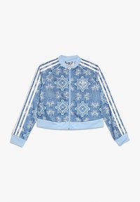 Chaqueta azul clara con cremallera y patrones geométricos blancos ornamentados, tres franjas blancas en las mangas, puños ribeteados y cuello.