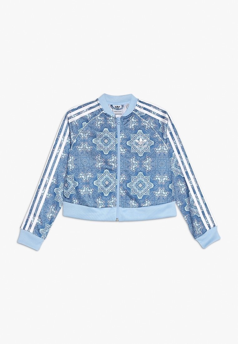 Chaqueta azul clara con cremallera y patrones geométricos blancos ornamentados, tres franjas blancas en las mangas, puños ribeteados y cuello.