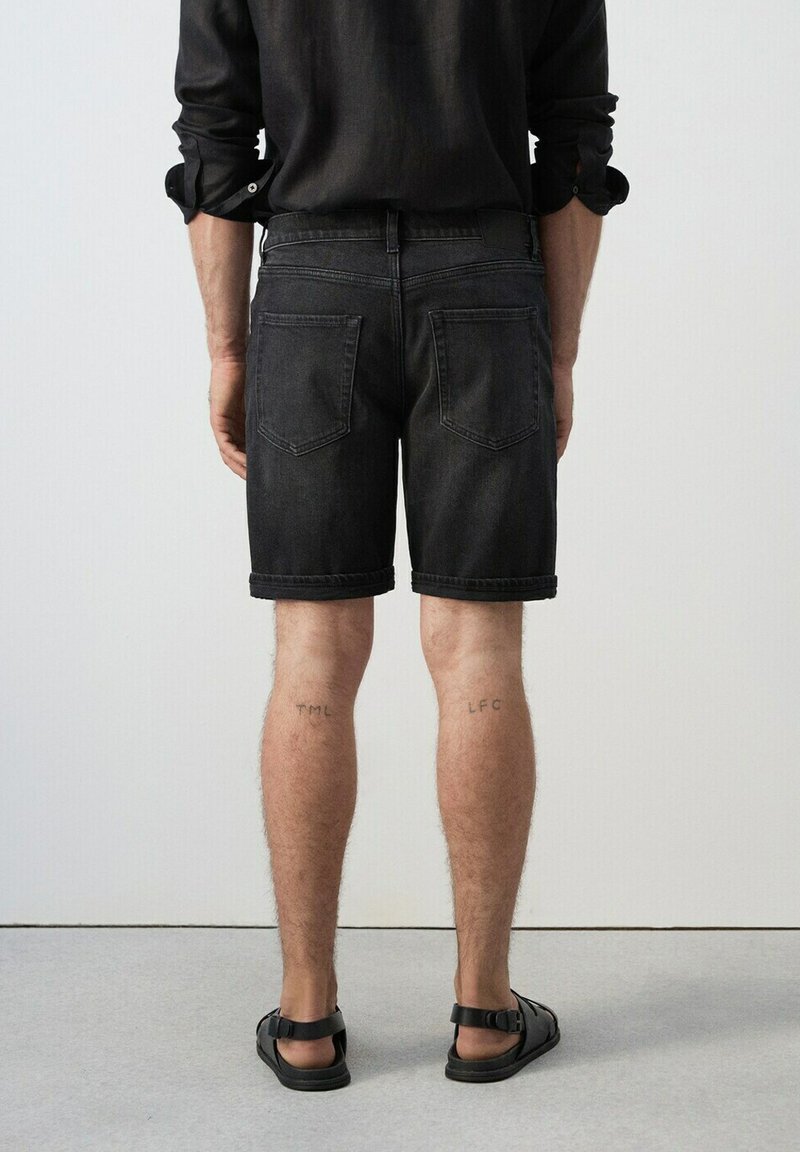 Homme portant un short en jean noir retroussé, une chemise noire à manches longues et des sandales noires, debout de dos sur un fond uni.