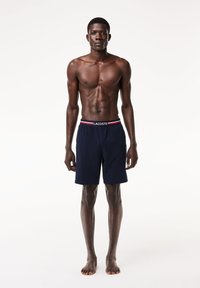 Pantaloni da mare blu navy con un tessuto morbido, vita elasticizzata con logo Lacoste, dettagli rossi e bianchi, e una vestibilità rilassata.