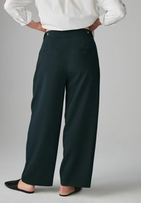 Dark green wide-leg pants made of smooth fabric. Features include a high waist, back pockets, and silver-tone hardware. Worn with black flats.