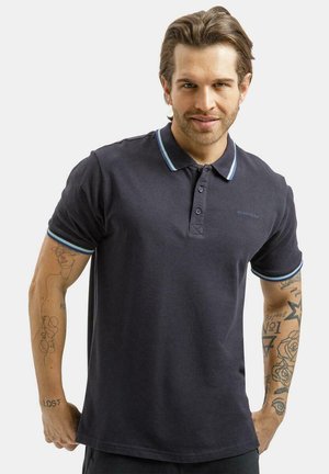 Mężczyzna o jasnobrązowych włosach, ubrany w ciemnogranatową koszulkę polo z niebieskimi i białymi lamówkami, pokazujący tatuaże na obu przedramionach.