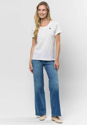 Noor blondide pikkade juustega noor naine, kes kannab valget lühikeste varrukatega särki, siniseid laiu pükse ja kreemjaid lamekontsaga jalanõusid, naeratab ja seisab.