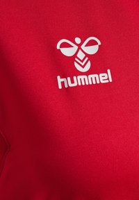 Tessuto rosso con un logo bianco che presenta un design astratto di un'ape e la parola "hummel" sottostante, caratterizzato da una texture liscia e da un colore vivace.