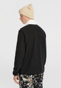 Zwarte lange mouw top met een witte kraag, gecombineerd met een broek met patronen in donkere en lichte vlekken. Beige gebreide muts op het hoofd.