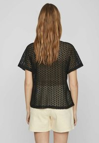Top de ganchillo negro con mangas cortas, que presenta un patrón geométrico y un corte holgado. Combinado con pantalones cortos de color crema, que muestran una superficie texturizada.