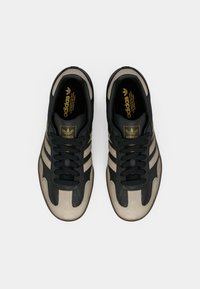 Μαύρα και μπεζ αθλητικά παπούτσια με υλικά δέρματος και καουτσούκ, με τρεις μαύρες ρίγες, στρογγυλά κορδόνια και χρυσή ετικέτα με το λογότυπο στη γλώσσα.