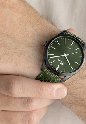 Orologio da polso con quadrante verde e cinturino in tessuto che segna le 4:18, tenuto da una mano mentre regola il cinturino su un polso dalla pelle chiara.