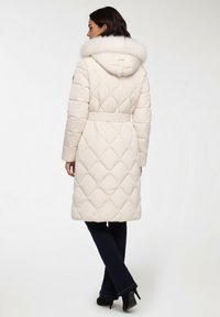 Cappotto lungo imbottito in beige chiaro con motivo trapuntato, cappuccio foderato in pelliccia e dettaglio della cintura in vita. Presenta maniche lunghe e una linea dritta.