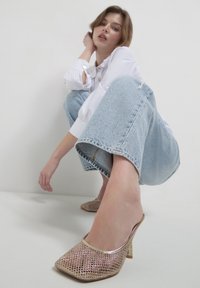 Donna in posizione accovacciata contro uno sfondo bianco, indossa jeans azzurri chiari, una camicia bianca con bottoni e scarpe con tacco alto in rete dorate.