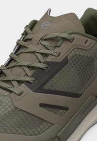 Κοντινό πλάνο αθλητικού παπουτσιού σε ελαιί πράσινο με υφή πλέγματος, επενδύσεις από σουέντ, κορδόνια με σχέδιο και μαλακό γιακά.