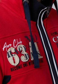 Rode rits hoodie met marineblauwe accenten, met geborduurde details: "Aero Club 63" en "Word Een met het Vliegtuig." Getextureerde stof.