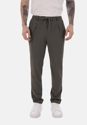 Uomo che indossa pantaloni casual grigio scuro con coulisse in vita e sneakers bianche, tatuaggi visibili sugli avambracci su sfondo bianco.