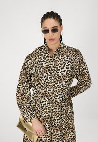 Beige, pruunide ja mustade leopardimustriga kleit, millel on pikad varrukad, nööbiga eesmine osa ja köitega vöökoht. Kaasas kuldne käekott.