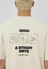 T-shirt en coton couleur crème avec des illustrations noires de notes autocollantes et le texte : "si ce n'est pas sur une NOTE AUTOCOLLANTE, ça n'existe pas."
