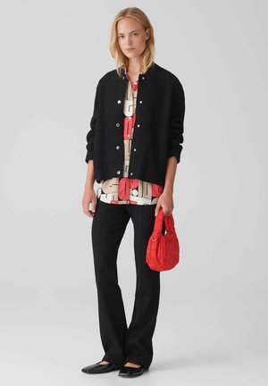 Zwarte cropped cardigan, gebloemde crèmekleurige en rode blouse, zwarte broek en een rode gewatteerde handtas. Vlakke zwarte schoenen maken de look compleet.
