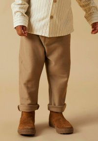 Pantaloni di cotone beige con orlo risvoltato, abbinati a stivali alla caviglia marroni. La camicia è bianca con righe verticali e bottoni in legno.