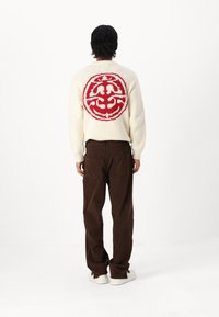Suéter de punto crema con un gran diseño circular rojo en la parte de atrás, combinado con pantalones de corduroy marrones y zapatillas blancas.