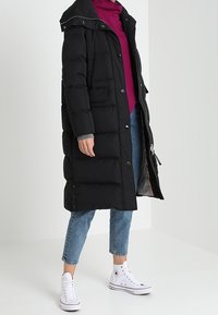 Abrigo acolchado negro con capucha, que presenta un cierre de cremallera y a presión, combinado con una camiseta burdeos y jeans de color azul claro, llevado con zapatillas blancas.