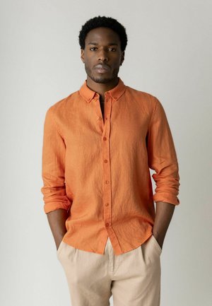 Man met kort krullend haar, gekleed in een oranje overhemd met opgerolde mouwen en beige broek, staand met handen in de zakken tegen een effen muur.