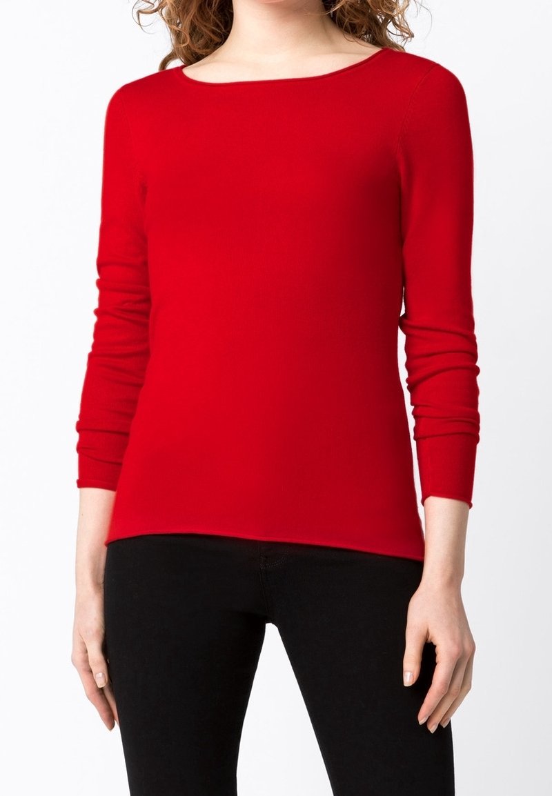 Femme portant un pull rouge ajusté à manches longues et un pantalon noir, debout devant un fond blanc uni.