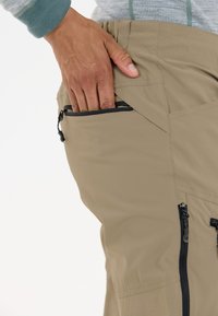 Un pantalon beige avec une finition texturée présente des poches zippées noires. La taille est élastique et une main est positionnée près de la poche latérale.