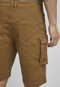Tan cotton cargo shorts with a side pocket, two back pockets, and stitching detail. The fabric has a smooth texture.