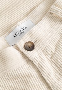 Kremno barvani kordurov blago z rjavim gumbom in belo oznako znamke, na kateri piše "LES DEUX copenhagen".