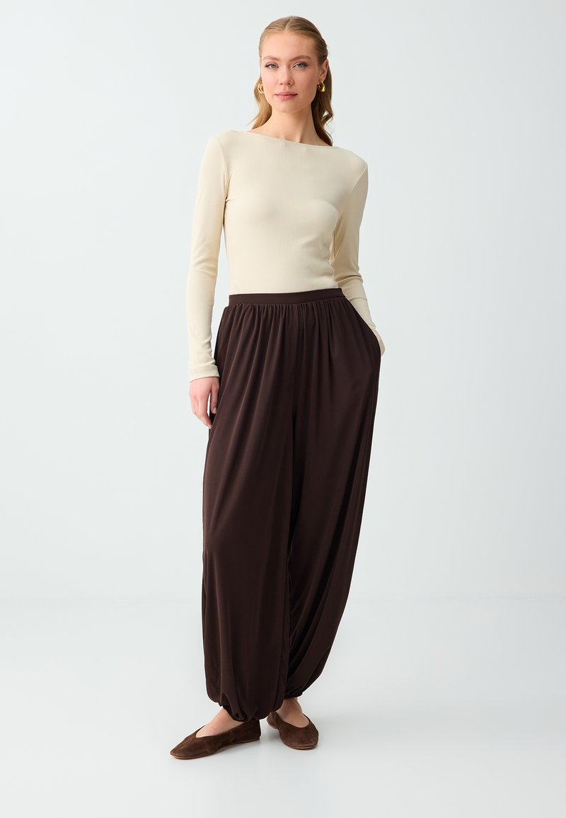 Femme debout, les mains dans les poches, portant un haut à manches longues crème, un pantalon ample marron foncé et des chaussures plates marron, sur un fond uni.
