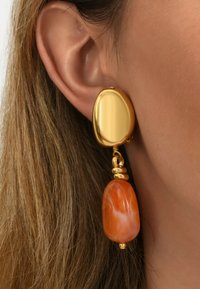 Boucle d'oreille en ton doré avec une forme lisse et irrégulière, ornée d'une pierre orange polie suspendue à l'oreille d'une personne.