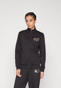 Giacca nera con zip realizzata in un tessuto morbido, dotata di una zip frontale, colletto alto e un design ricamato decorativo sul petto e sulla cintura.