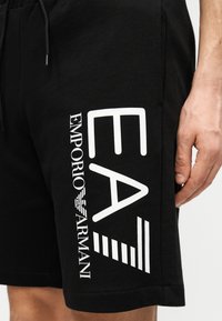 Μαύρα βαμβακερά σορτς με μεγάλο λευκό γραφικό που περιλαμβάνει το "EA" και το κείμενο "Emporio Armani", συμπληρωμένα από λωρίδες σχεδιαστικών λεπτομερειών.