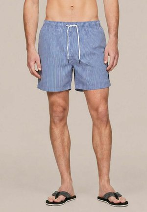 Man die blauwe en witte gestreepte zwembroek draagt met een witte trekkoord en zwarte teenslippers, staand tegen een effen achtergrond.