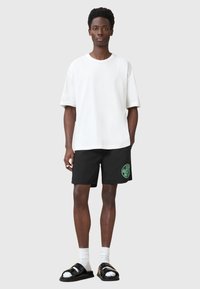 Camiseta blanca oversized, pantalones cortos negros con detalle del logo en verde, sandalias negras, calcetines blancos. Conjunto casual sobre un fondo neutro.