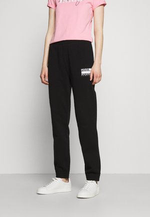 Person trägt schwarze Sweatpants mit Hugo-Logo, pinkes kurzärmeliges Shirt und weiße Sneaker, steht vor einem einfarbigen hellen Hintergrund.