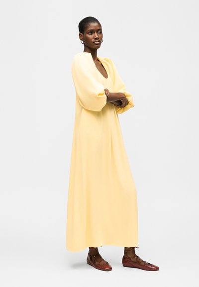 Γυναίκα με κοντά μαλλιά που φοράει μακρύ, άνετο κίτρινο φόρεμα και καφέ δερμάτινα παπούτσια με κορδόνια, στέκεται με σταυρωμένα χέρια, κοιτάζοντας ελαφρώς προς τα δεξιά.