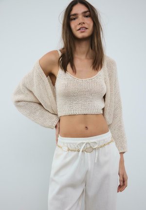 Mladá žena v béžovom pletenom krátkom topánku, zodpovedajúcom kardigáne padajúcom z jedného ramena, bielych nohaviciach a zlatom reťazovom opasku.