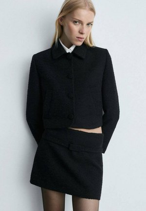 Black cropped jacket in textured fabric with a rounded collar, front buttons, and side pockets, paired with a matching skirt with a belt.