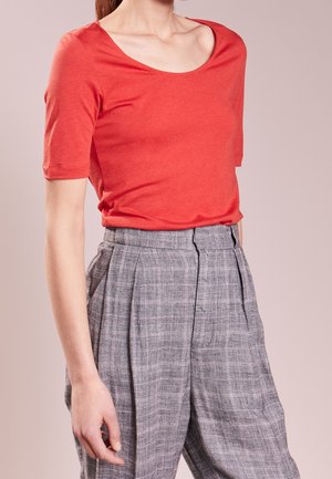 Mujer con una blusa roja de manga corta con escote asimétrico y pantalones gris de cuadros de talle alto sobre un fondo liso.