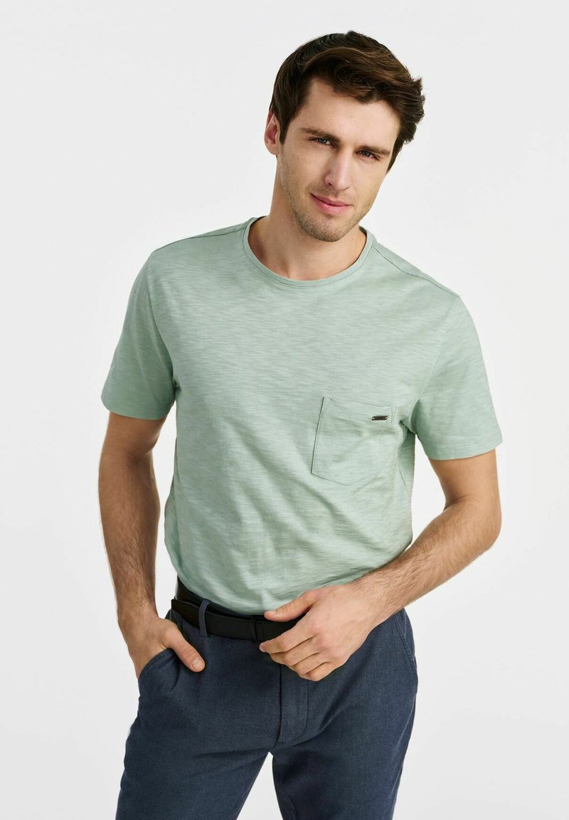 Jasnozielona koszulka z krótkim rękawem i okrągłym dekoltem, z kieszonką z przodu i delikatną teksturą. Noszona z ciemnymi spodniami.