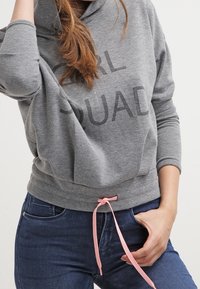 Sudadera gris con el texto "GIRL SQUAD", cordón con acentos en rosa y corte relajado. Combinada con jeans oscuros.
