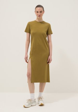 Vrouw staat met het gezicht naar voren, draagt een olijfgroene midi-jurk met korte mouwen en een hoge split op de dij, en witte sneakers met sokken.