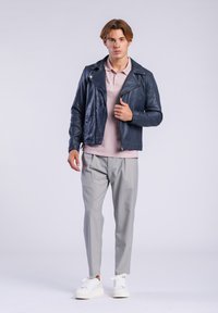 Chaqueta de cuero de motociclista navy con cremalleras, polo rosa, pantalones rectos gris claro y zapatillas blancas. Hombre en un fondo neutral.