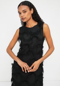 Mujer con cabello rizado vistiendo un vestido negro sin mangas con patrones de flecos circulares texturizados, manos suavemente entrelazadas en la cintura, fondo neutro.