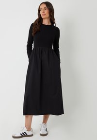 Abito lungo nero con maniche lunghe, dal taglio aderente e vita arricciata, con tasche laterali. Abbinato a sneakers bianche con strisce nere.