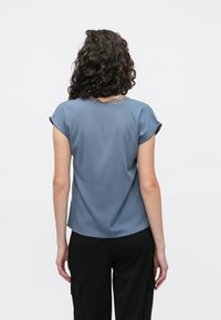 Sininen lyhythihainen paita, joka on valmistettu pehmeästä kankaasta ja jossa on pitsiyksityiskohta kaula-aukossa. Takana paita on sileä ja hieman väljä.