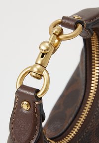 Dettaglio della tracolla di una borsa in pelle marrone con un clip in metallo dorato e una cerniera. La pelle presenta una finitura texturizzata con un motivo sottile.
