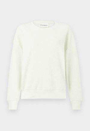 Jasnobeżowa teksturowana bluza, ciemne jeansy z szerokimi nogawkami, zielone zamszowe buty oraz mała czarna skórzana torba. Modelka stoi ze swobodnie opuszczonymi ramionami.